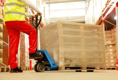 İşçi ambalajlı ahşap paletleri depoda manuel forklift ile hareket ettiriyor.