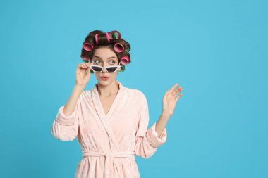 Emotional young woman in bathrobe with hair curlers and sunglasses on light blue background