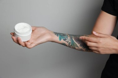 Woman applying cream on her arm with tattoos against light grey background, closeup