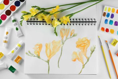 Painting of freesias in sketchbook, flowers and art supplies on white background, top view
