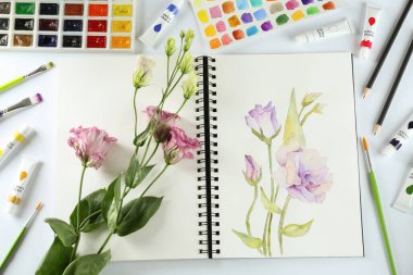 Painting of eustomas in sketchbook, flowers and art supplies on white background, top view