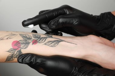 Worker in gloves applying cream on woman's arm with tattoo against light background, closeup