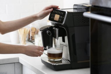 Woman using modern espresso machine for making coffee with milk in kitchen, closeup