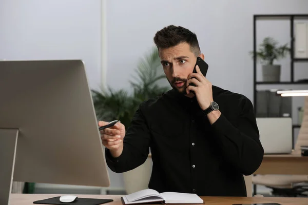 オフィスのテーブルでコンピューターと仕事をしながら電話で話す男 — ストック写真