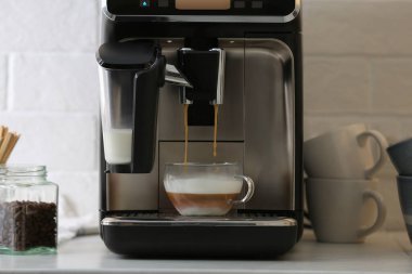 Modern espresso makinesi mutfaktaki beyaz tezgahın üzerine sütlü cam bardağa kahve dolduruyor.