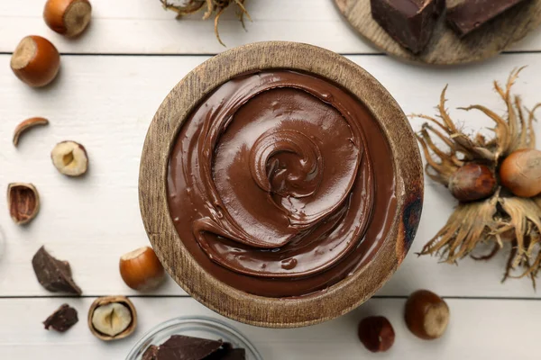 Bowl of tasty chocolate paste with hazelnuts on white wooden table, flat lay