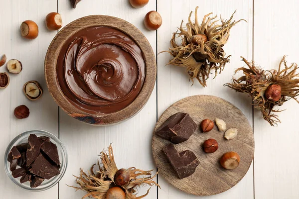 Bowl of tasty chocolate paste with hazelnuts on white wooden table, flat lay