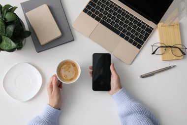 Beyaz ofis masasında kahve ve akıllı telefon kullanan bir kadının en iyi görüntüsü.