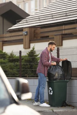 Adam çöp torbasını geri dönüşüm kutusuna koyuyor.