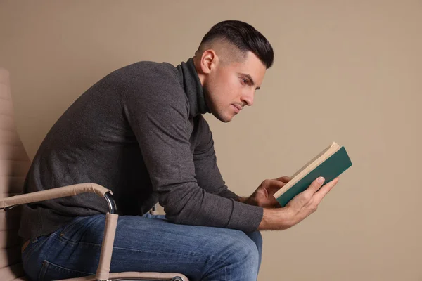 Człowiek Słabą Postawą Czytania Książki Siedząc Krześle Beżowym Tle — Zdjęcie stockowe