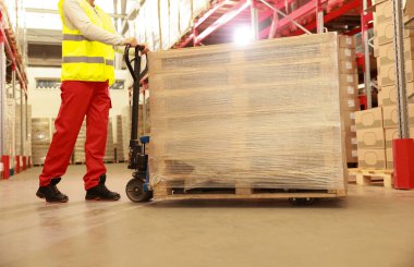 İşçi ambalajlı ahşap paletleri depoda manuel forklift ile hareket ettiriyor.