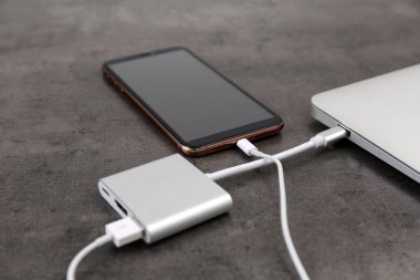 Gri masadaki elektrik bankasından şarj olan modern akıllı telefon ve laptop, yakın plan.