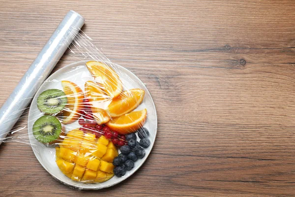 Plate of fresh fruits and berries with plastic food wrap on wooden table, flat lay. Space for text