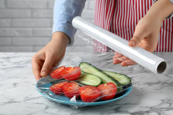 Woman putting plastic food wrap over plate of fresh vegetables at white marble table indoors, closeup