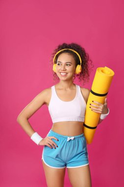 Pembe arka planda yoga minderi ve kulaklıkları olan güzel Afrikalı Amerikalı kadın.