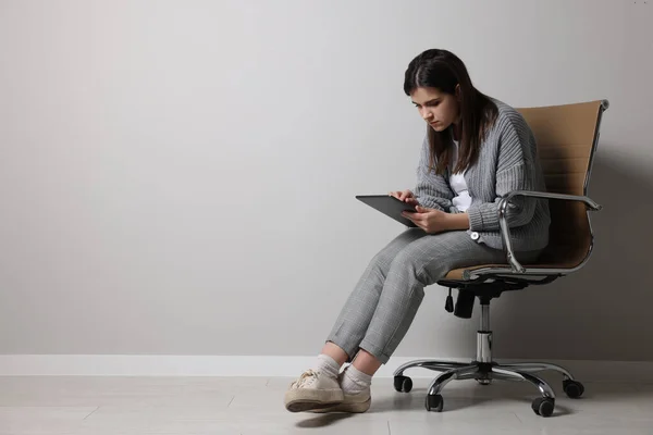 Young woman with bad posture using tablet while sitting on chair near grey wall. Space for text