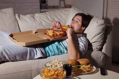 Şişman adam evdeki kanepede pizza yiyor.