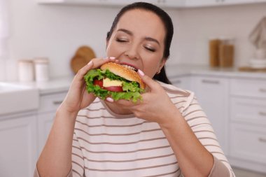 Kilolu bir kadın mutfakta hamburger yiyor.