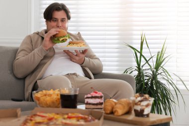 Kilolu bir adam evdeki kanepede lezzetli bir burger yiyor.