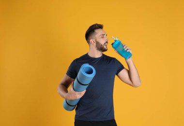 Yoga minderli yakışıklı adam sarı arka planda su içiyor.