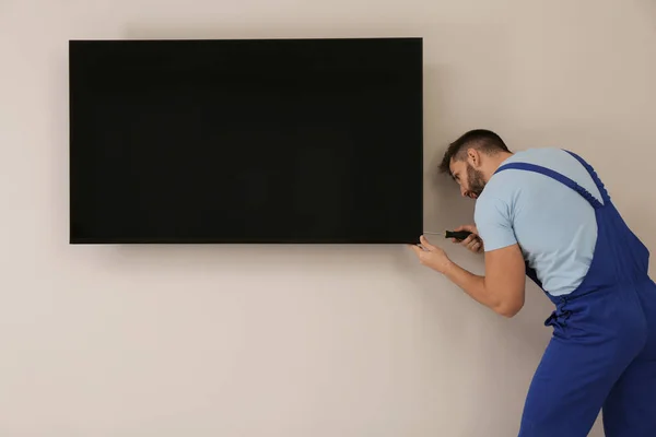 Professional technician with screwdriver installing modern flat screen TV on wall indoors