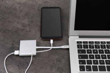 Gri masadaki elektrik bankasından şarj olan modern akıllı telefon ve laptop, yakın plan.