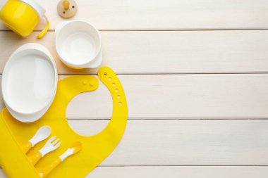 Beyaz ahşap zemin üzerinde silikon bebek önlüğü ve plastik bulaşık takımı olan düz bir kompozisyon. Metin için boşluk