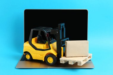 Dizüstü bilgisayar, tahta paletli oyuncak forklift ve açık mavi arka planda kutu.