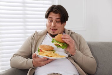 Evdeki kanepede bir tabak hamburger ve patates kızartması olan şişman bir adam.