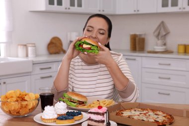 Kilolu bir kadın mutfakta hamburger yiyor.