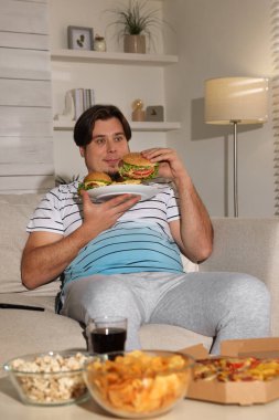Evdeki kanepede bir tabak hamburger ve patates kızartması olan şişman bir adam.