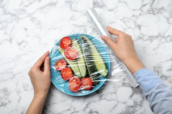 Woman putting plastic food wrap over plate of fresh vegetables at white marble table, top view