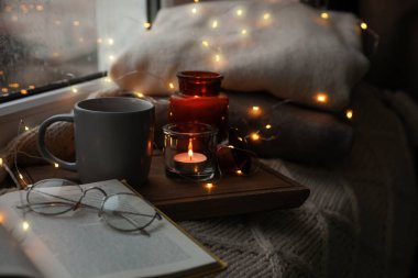 Bir fincan sıcak içecek, açık kitap ve pencerenin yanındaki örgü battaniyenin üzerinde Noel ışıkları. Sıcak mevsim