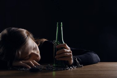 Alkol bağımlılığı. Bir şişe birayla zincirlenmiş, siyah arka planda tahta masada uyuyan bir kadın.