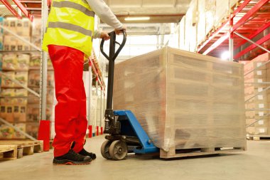 İşçi ambalajlı ahşap paletleri depoda manuel forklift ile hareket ettiriyor.