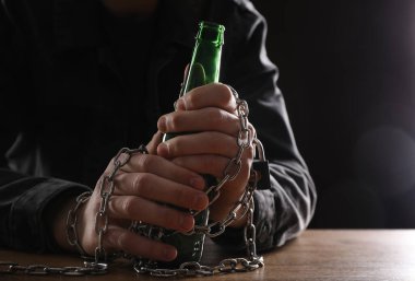 Alkol bağımlılığı. Ahşap masaya bira şişesiyle zincirlenmiş bir adam, yakın plan.