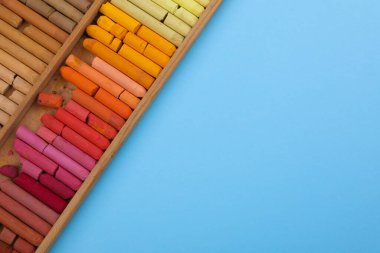 Açık mavi arkaplandaki ahşap kutuda yumuşak pastel renkler seti, üst görünümü metin için alan. Çizim malzemesi