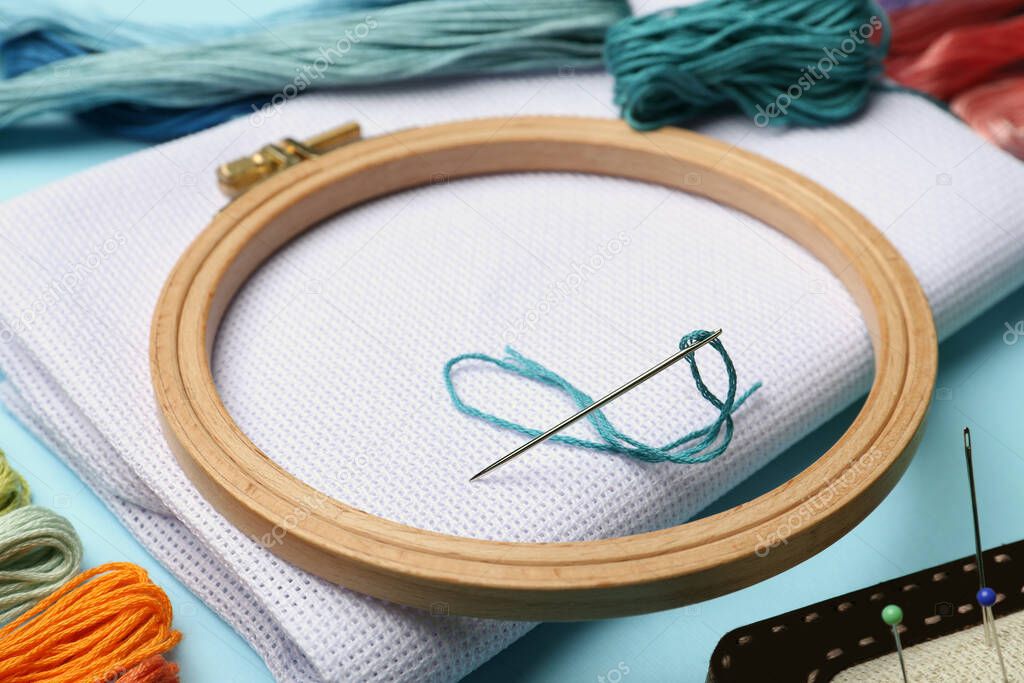 Embroidery hoop, different threads, fabric and needles on light blue background, closeup