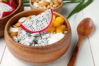Bir kase granola, pitahaya, mango, badem ve yoğurt beyaz ahşap masada.