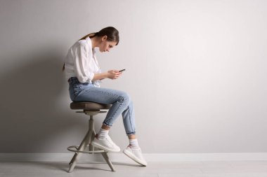 Akıllı telefon kullanan kötü duruşu olan bir kadın açık gri duvarın yanındaki taburede otururken, mesaj için boş yer