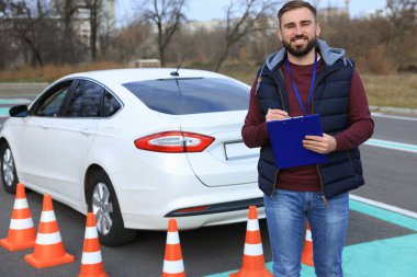 Dışarıdaki arabanın yanında panosu olan bir eğitmen. Ehliyet sınavı
