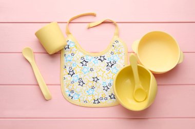 Salya akıtan bebek önlüğü ve pembe ahşap arka planda plastik bulaşık takımı ile düz bir kompozisyon.