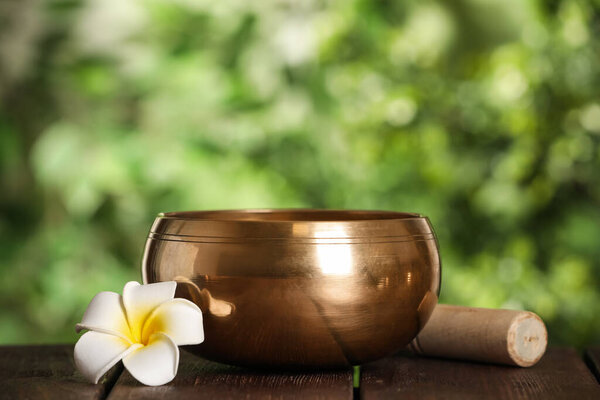 Golden singing bowl, mallet and flower on wooden table outdoors