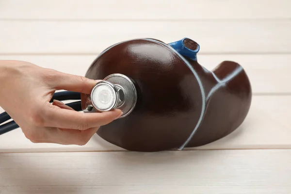 Doctor with stethoscope examining liver model at white wooden table, closeup