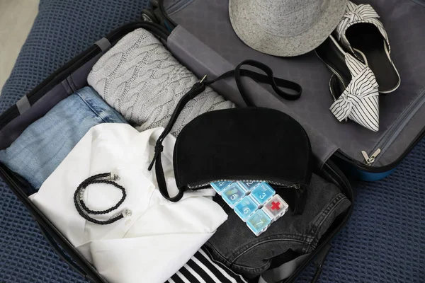 Open suitcase with packed clothes, accessories and pill box on blue blanket, above view