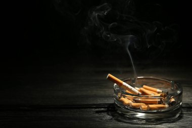 Siyah arka plana karşı koyu ahşap masada cam kül tablasında sigara yakmak. Metin için boşluk