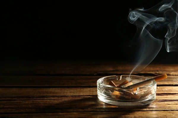 在黑色背景的木制桌子上的玻璃烟灰缸里燃烧着香烟 案文的篇幅 — 图库照片