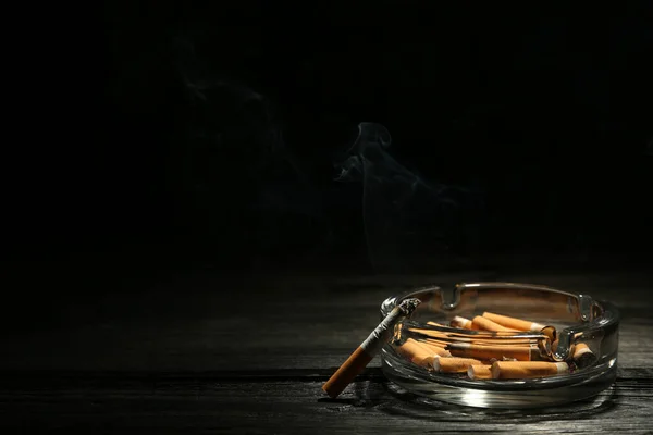 玻璃烟灰缸 黑色背景的深色木制桌子上有烟蒂和燃烧的香烟 案文的篇幅 — 图库照片