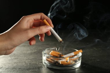 Gri masadaki cam kül tablasının üzerinde sigara içen kadın siyah arka planda, yakın plan.