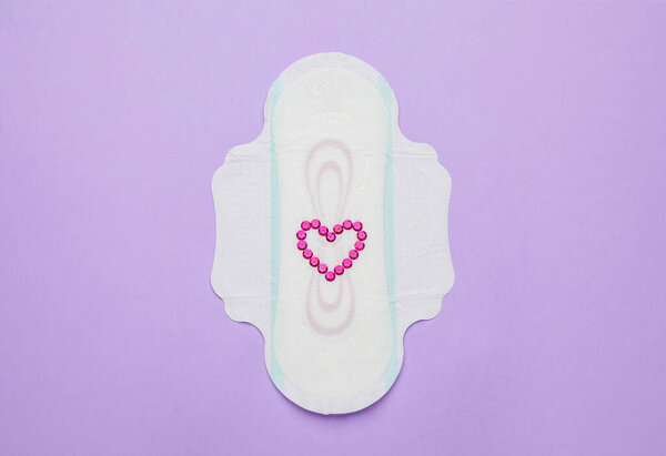 Menstrual pad with heart made of pink blins and on violet background, top view